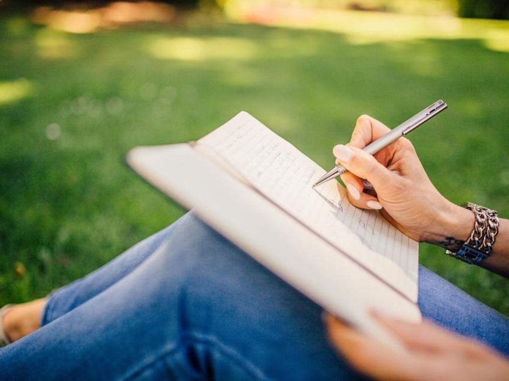 A person writing notes on a pad, sitting outside on the grass
