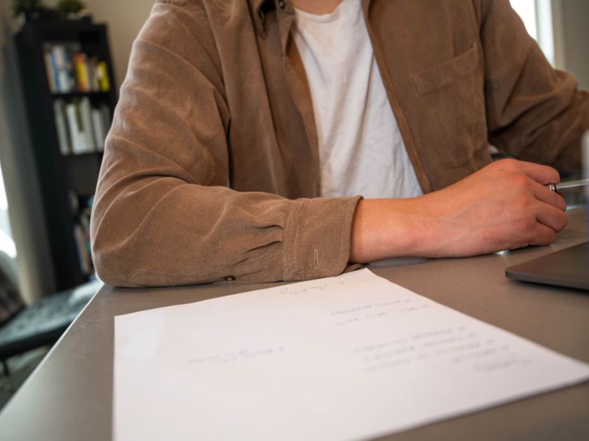 A person at a desk with a document in front of them and a pen in hand, a bookshelf behind, seen from the shoulders down