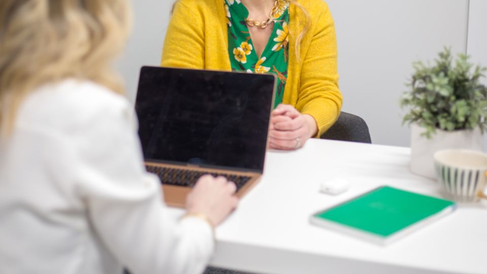 Two people across a white table, one with a laptop open, a green notebook between them, both heads out of frame