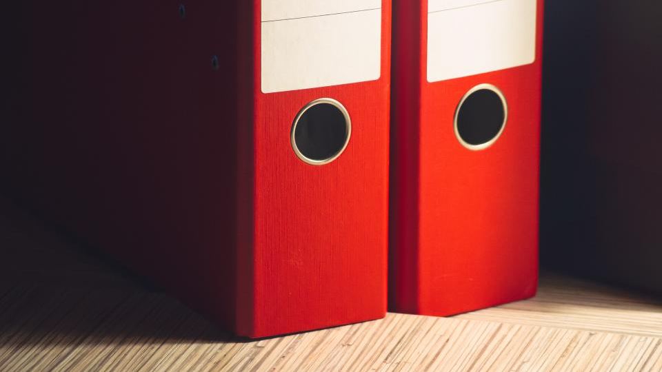 Two red lever-arch folders standing on a wooden shelf, their spines unlabelled
