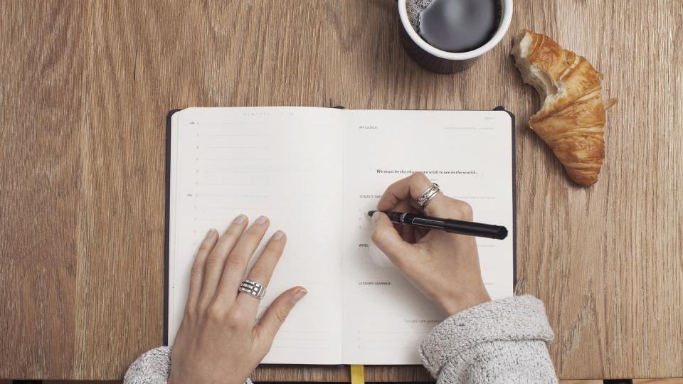 Hands writing in an open notebook on a wooden table beside a coffee