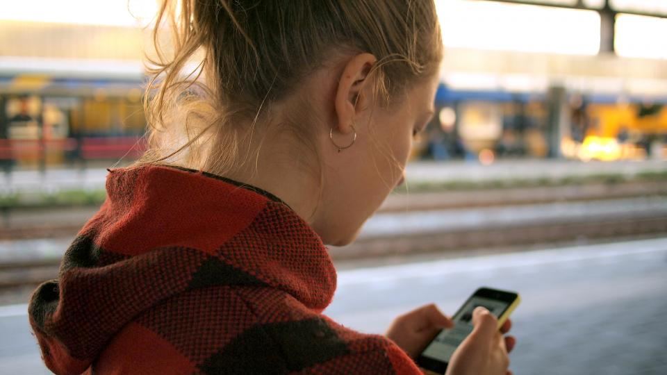 A person seen from behind on a railway platform, reading something on their phone, a train blurred beyond them