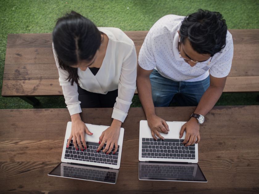 Two people side by side at a wooden bench, each typing on a laptop, seen from directly above